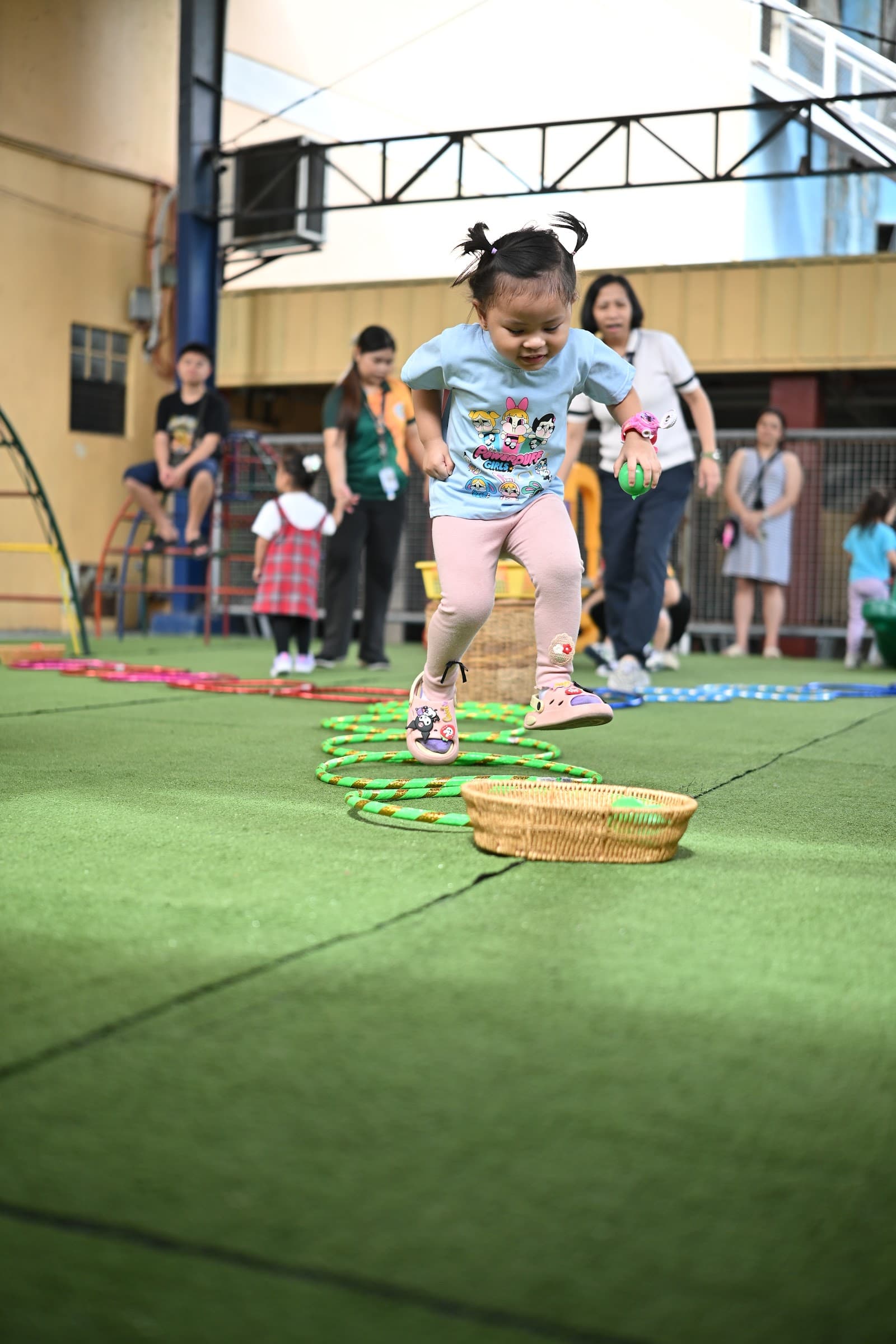 A toddler leaping through an agility ring course, laughing, on the school's outdoor play area