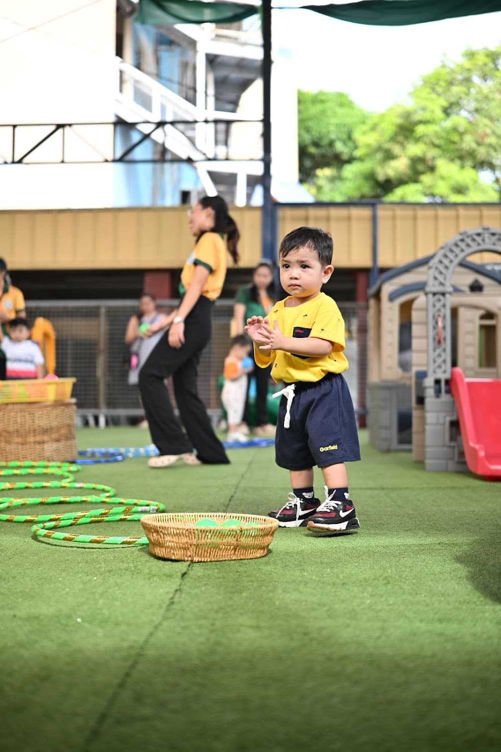 A toddler standing on the outdoor play turf near the playhouse